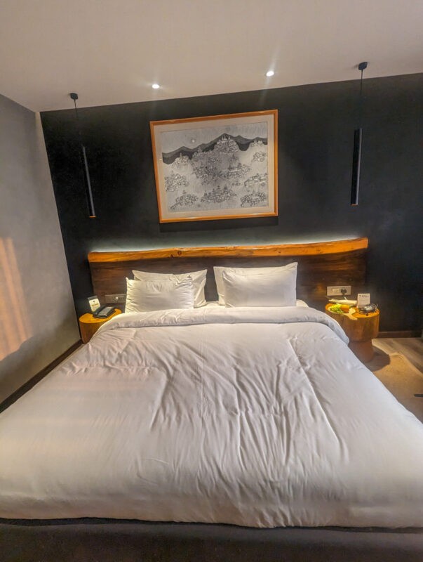 A modern hotel room featuring a large bed with white linens and a large, framed map above the headboard, against a dark gray wall.