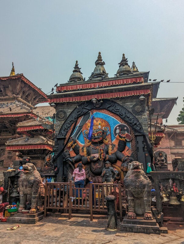 A dramatic temple entrance in Nepal, guarded by large, fierce-looking mythological creature statues, with locals and tourists milling about the area.