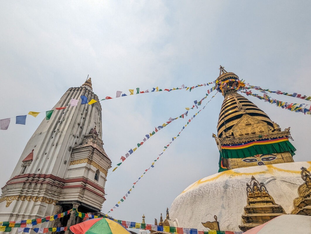 A magnificent view of a white stupa adorned with gold, surrounded by colorful prayer flags against a partially cloudy sky.