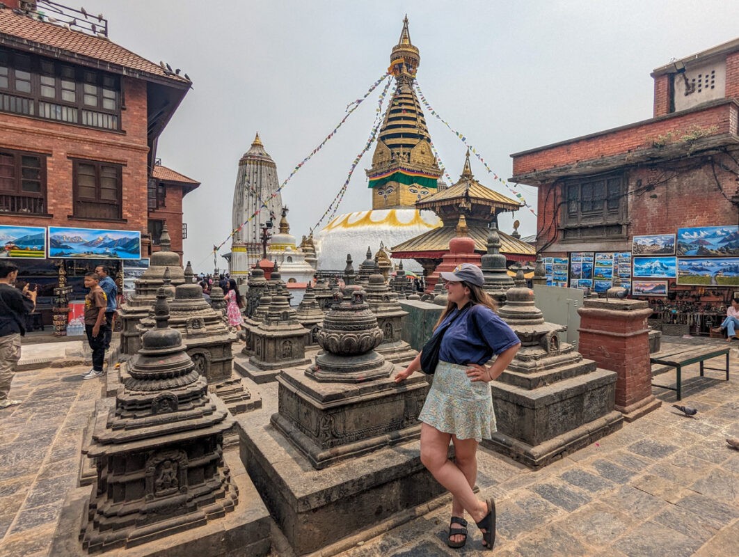 A bustling street scene in Kathmandu, Nepal, with a young woman standing in the foreground. Traditional Nepalese architecture surrounds the area, including intricate temples and colorful prayer flags stretching across the street.