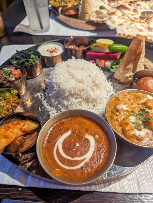 A vibrant array of Indian dishes arranged on a wooden table, featuring a mound of fluffy white rice surrounded by various curries including a rich, creamy dal makhani with a swirl of cream, palak paneer, and a chickpea curry. A side of naan bread, fresh vegetables, and small bowls of condiments complete the meal.