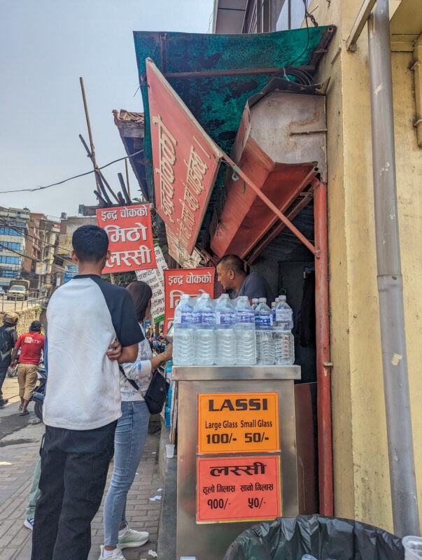 Busy street scene showing a vendor selling bottled water and lassi with signage in Nepali and English, next to a makeshift wooden structure.