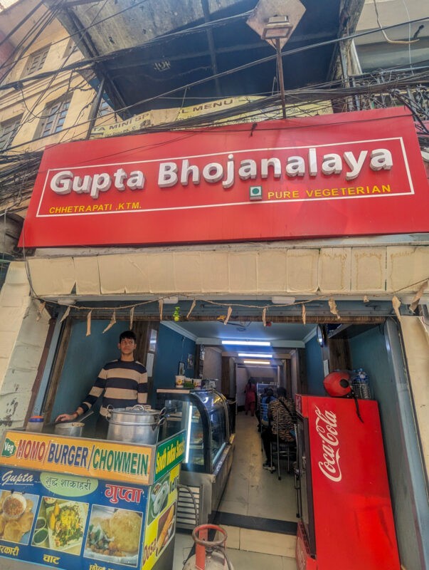 Entrance to Gupta Bhojanalaya, a narrow vegetarian restaurant with a red and white signboard, featuring a man standing behind a food counter.