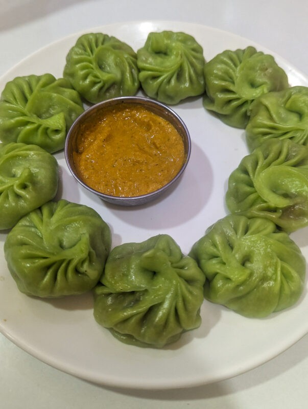 A white plate containing a circle of green momos with a small bowl of orange dipping sauce in the center