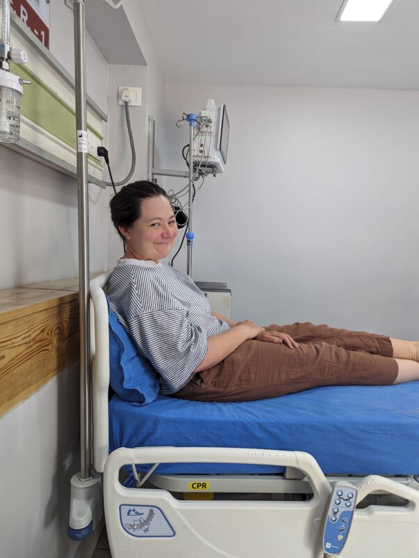 The image shows a smiling woman sitting on a hospital bed. She is dressed in a striped, blue and white button-down shirt and brown pants, reclining slightly against a blue bedsheet. To her right are medical devices including an IV stand and monitors, indicating she is in a medical or hospital setting. The room is brightly lit, with white walls and a small window near the ceiling.