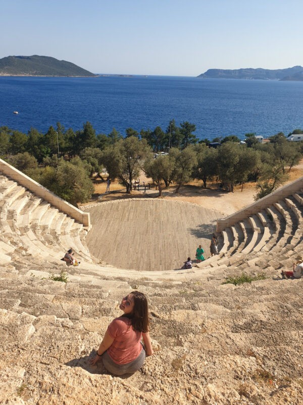 A woman sitting on ancient amphitheater steps, overlooking a scenic view of the blue sea and distant hills.