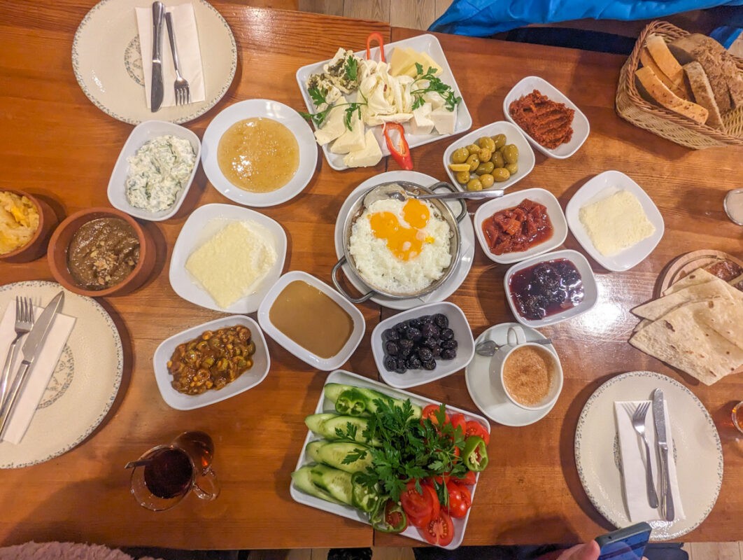 A traditional Turkish breakfast spread on a wooden table, featuring various dishes including eggs, olives, cheese, vegetables, and bread.