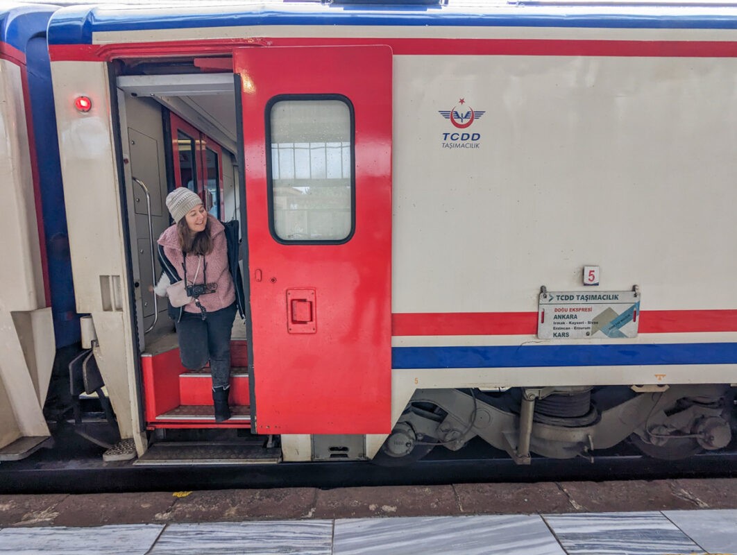 A woman stepping out of a red and white train carriage, bundled up in winter clothing, holding a camera.