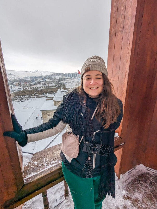 A woman in winter clothing, smiling while standing in a wooden balcony with a snowy cityscape in the background.