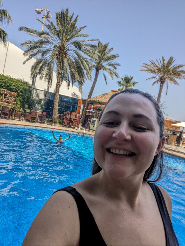A woman smiling at the camera while taking a selfie at an outdoor swimming pool, with palm trees and a person swimming in the background.