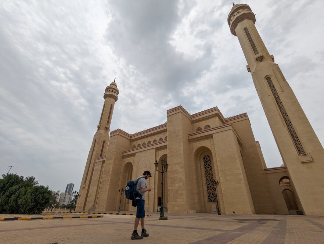 A person standing in front of a grand mosque with two tall minarets, looking at their phone under a cloudy sky.