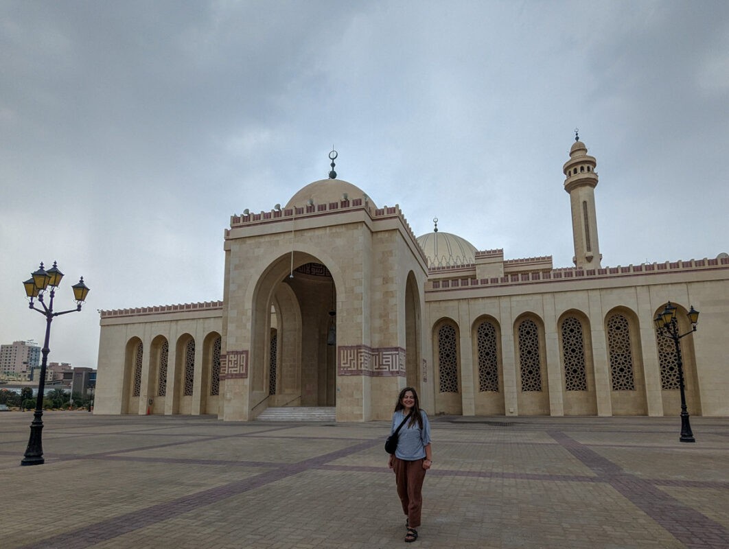 A woman standing in front of a large mosque with intricate architecture and multiple arches, under a cloudy sky.