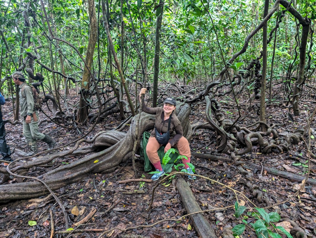 A person sitting on a large twisted tree root in a dense jungle, smiling at the camera while another person stands in the background.