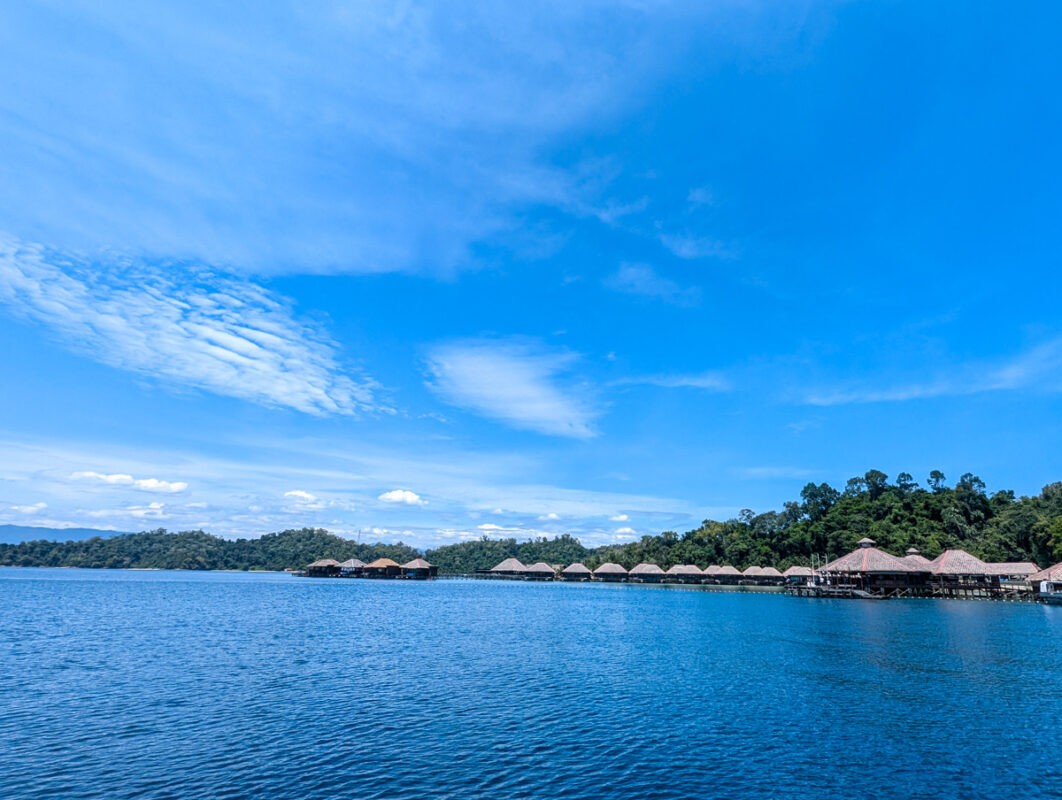 A row of overwater bungalows under a bright blue sky, with the ocean in the foreground.