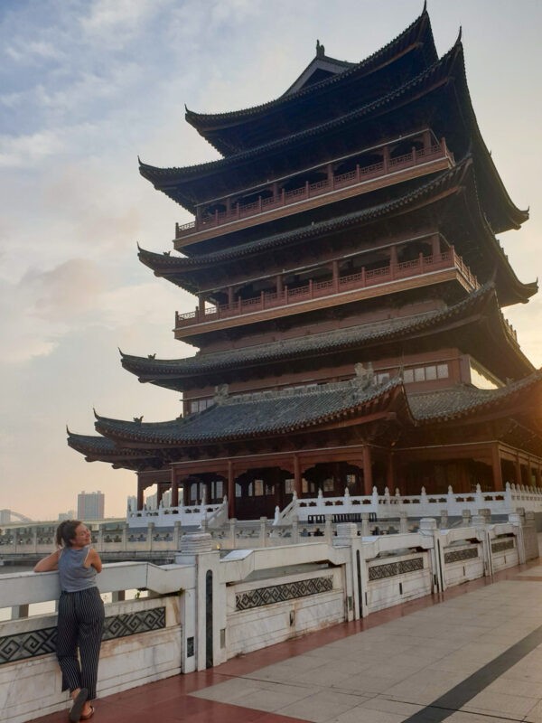 A woman leans against a railing, gazing up at a tall, traditional Chinese pagoda-style building with multiple tiers, set against a sky with soft clouds and warm light from the setting sun.