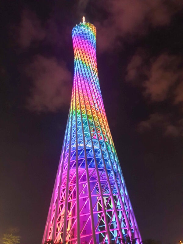 A vibrant, illuminated tower glows with a rainbow of colors against the dark night sky, showcasing a modern architectural design with a twisting form.