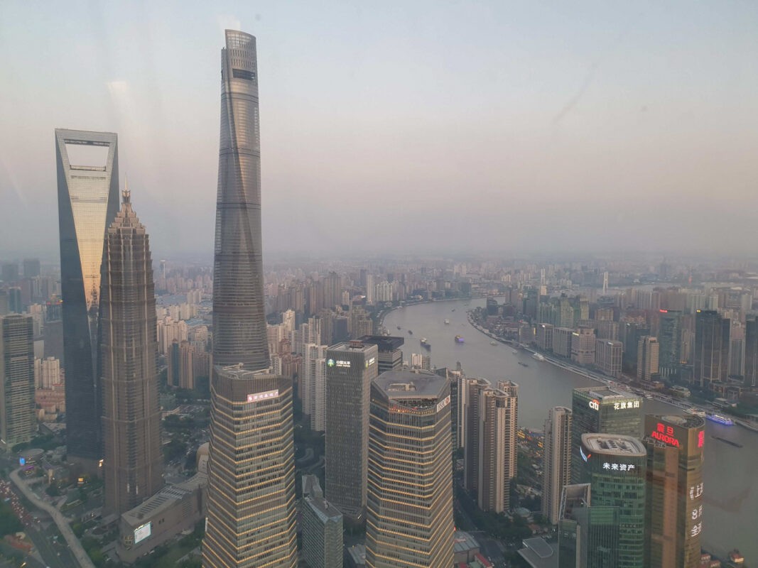 A panoramic cityscape of Shanghai at dusk, featuring iconic skyscrapers, including the Shanghai Tower, World Financial Center, and Jin Mao Tower, with the Huangpu River winding through the city.