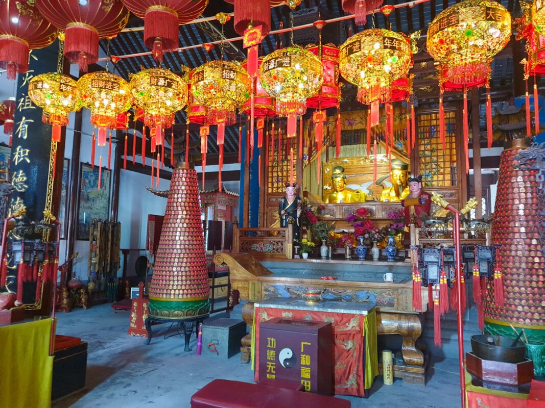 The interior of a traditional Chinese temple, richly decorated with golden statues, intricate woodwork, and numerous red and gold lanterns hanging from the ceiling.