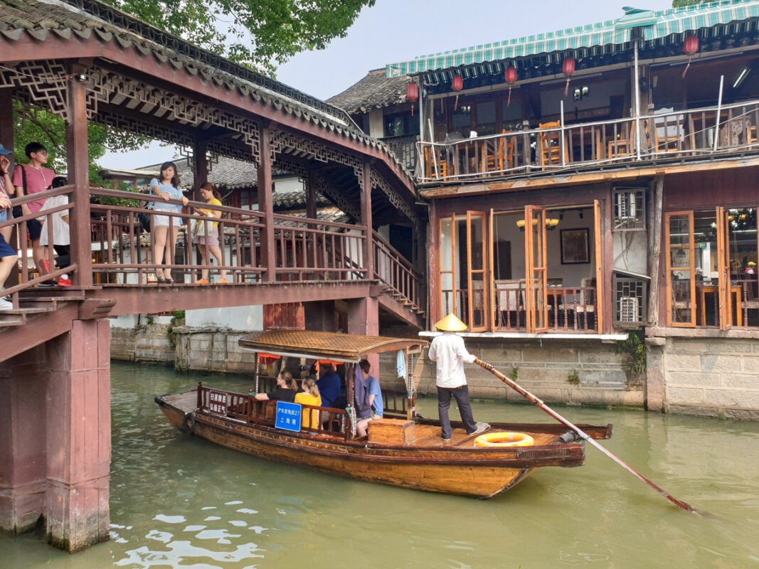 A traditional wooden boat with a gondolier wearing a conical hat navigates through a narrow canal flanked by old-style Chinese buildings and a small wooden bridge, with tourists observing from above.