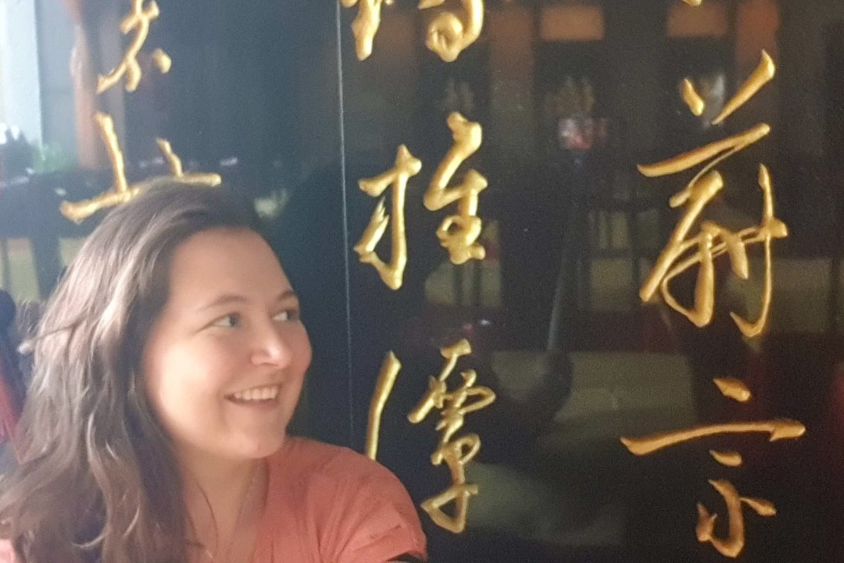 A woman smiles while standing in front of a black wall with large, gold Chinese calligraphy characters prominently displayed behind her.