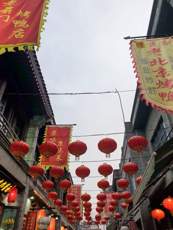 Red lanterns hang in rows above a busy alley lined with traditional Chinese shops, with large red and yellow banners featuring Chinese characters adding to the festive atmosphere.