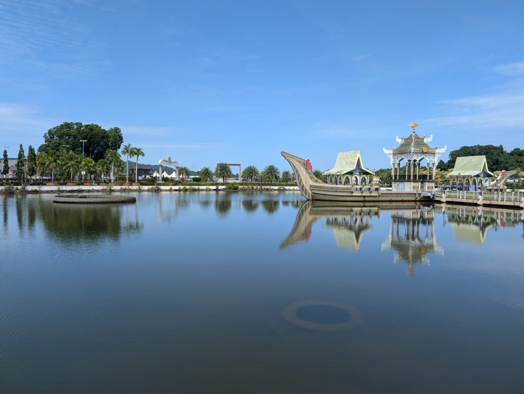 A serene lake scene with a floating pavilion and boat-shaped structure, reflecting on the still water under a clear blue sky. The shore is lined with palm trees and other greenery, with a backdrop of small buildings and a blue sky.