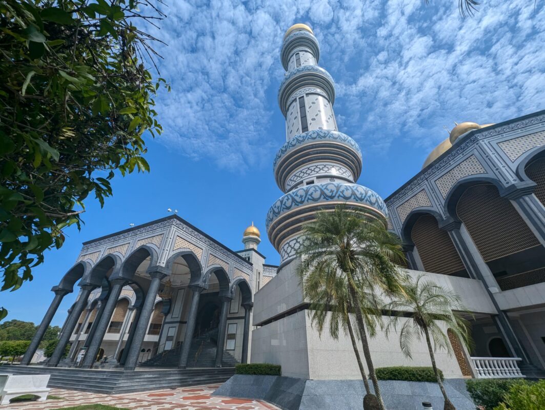 A wide shot of a mosque complex with multiple golden domes and tall minarets, surrounded by trees and an expansive parking area. A covered walkway leads towards the mosque, all under a bright blue sky.