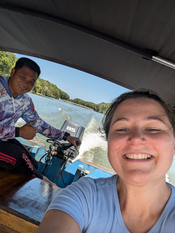 A selfie taken on a boat with two people smiling at the camera. The boat is moving through a river, and the background shows a lush green forest and the water’s wake.