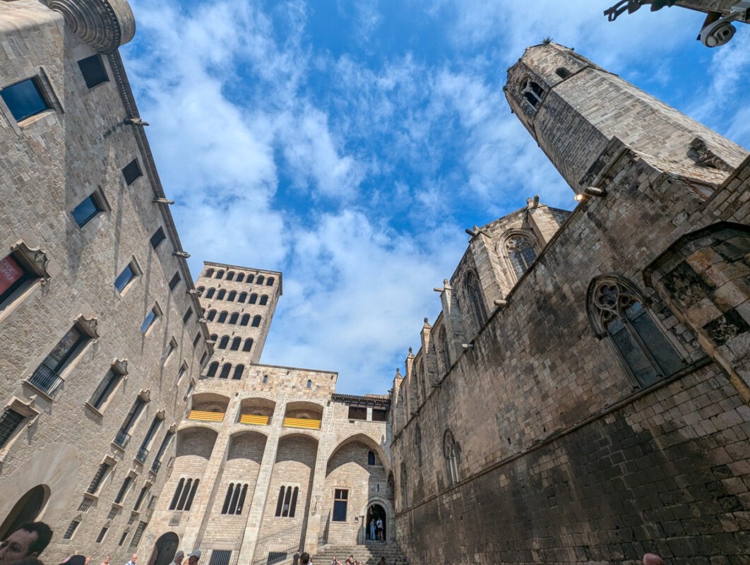 An upward view of historic stone buildings with arched windows and a large tower set against a bright blue sky with scattered clouds. The stone structures feature intricate Gothic architecture, including pointed arches and detailed carvings.