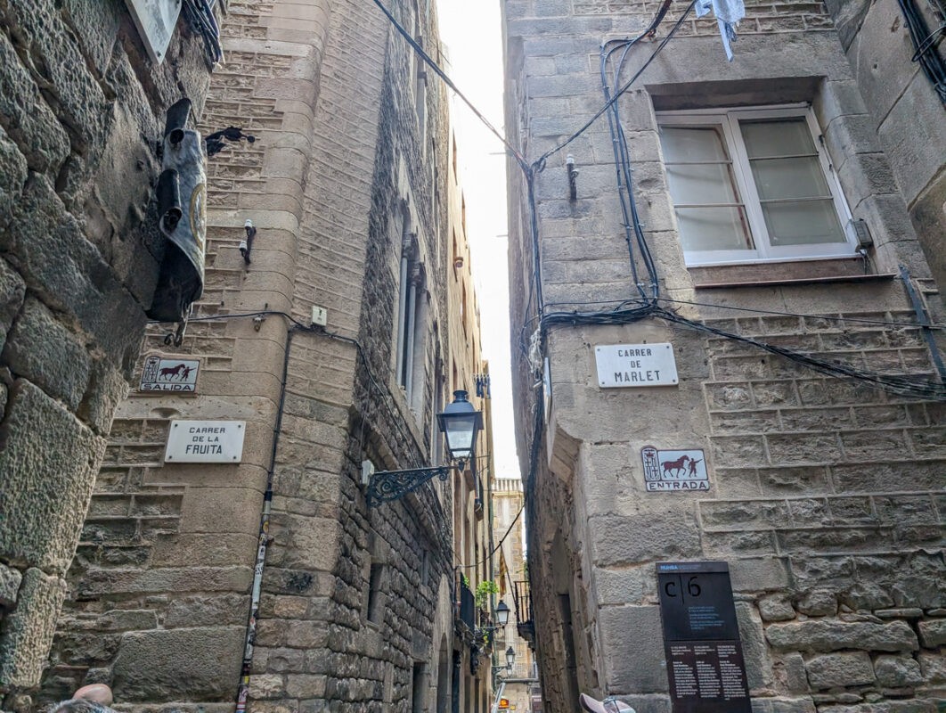 A narrow alleyway in an old town lined with stone buildings. Signs on the walls indicate street names such as "Carrer de la Fruita" and "Carrer de Marlet," with rustic street lights and exposed wiring overhead.