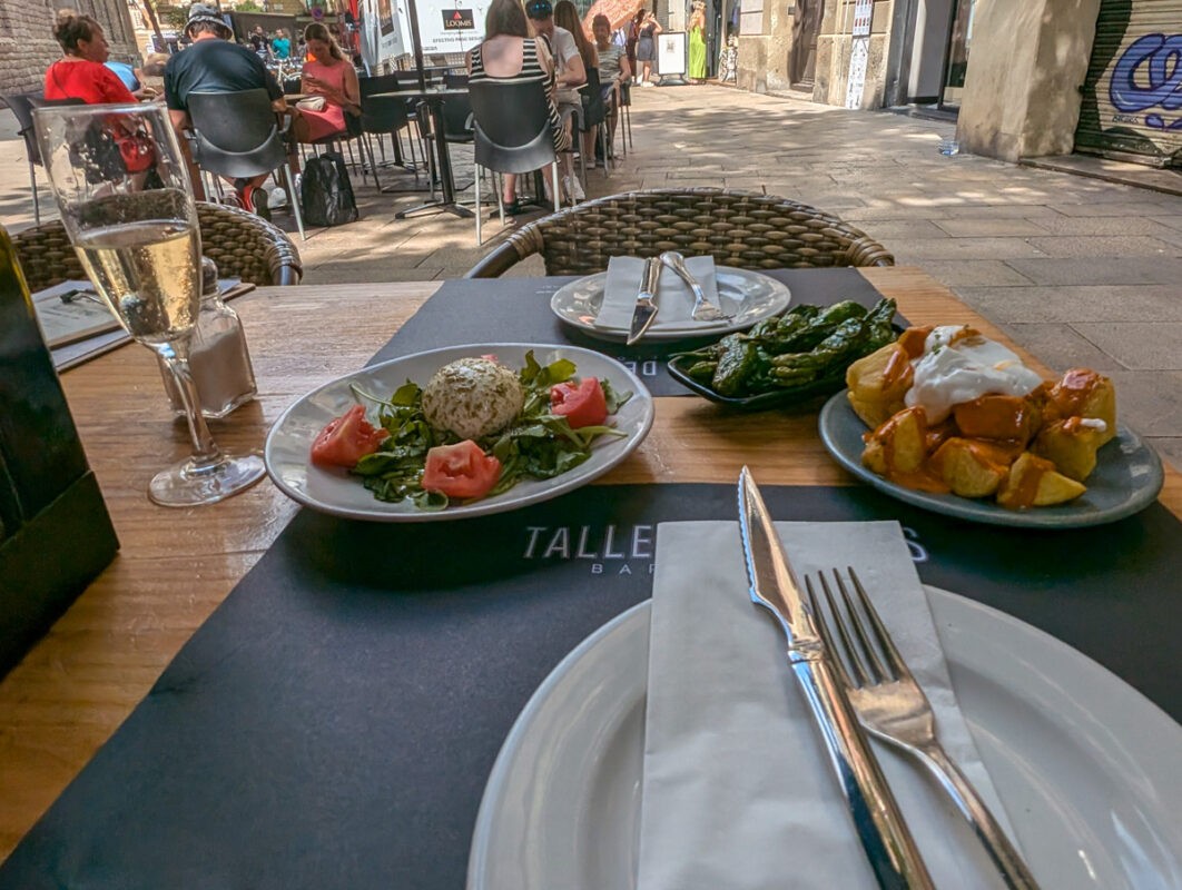 A table at an outdoor restaurant with plates of Spanish tapas, including Pimientos de Padrón, patatas bravas with a sauce, and a salad with tomatoes and a ball of mozzarella. A glass of champagne sits to the left of the dishes.