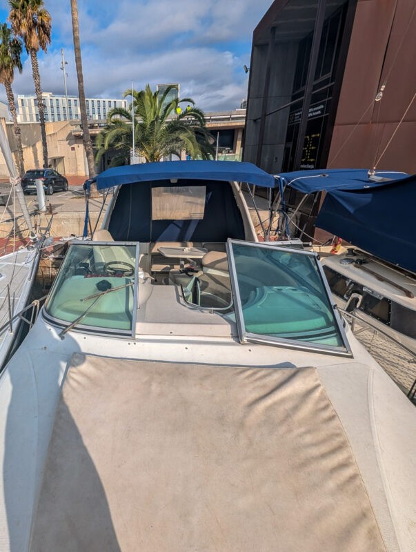 The front of a small boat docked in a marina, showing its open sun deck with a blue canopy. In the background are palm trees and a modern building, with a partly cloudy sky overhead.