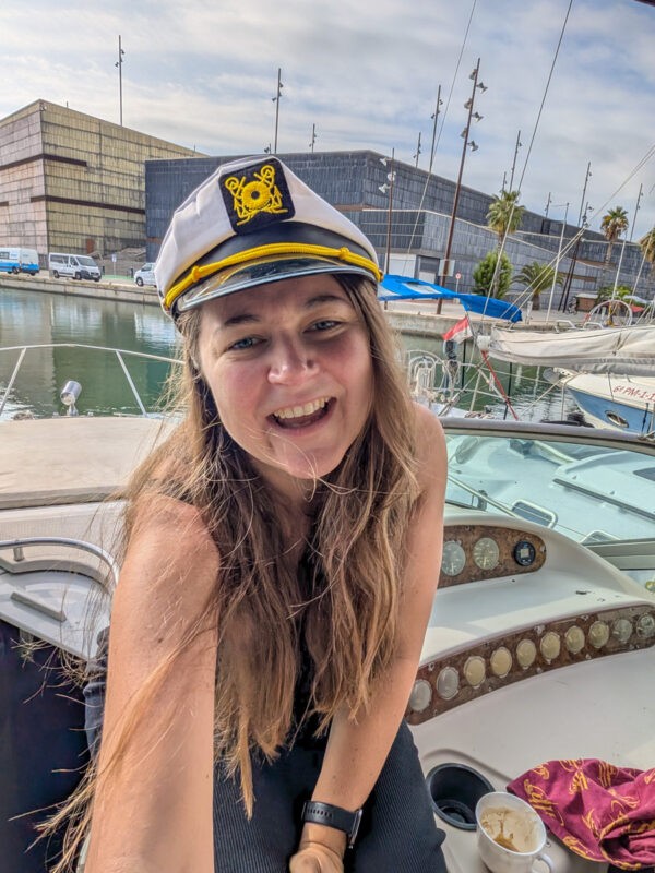 A woman smiling in a selfie while sitting on a boat. She is wearing a white captain’s hat with a navy and gold emblem. The marina is visible in the background, along with several other docked boats and modern buildings.