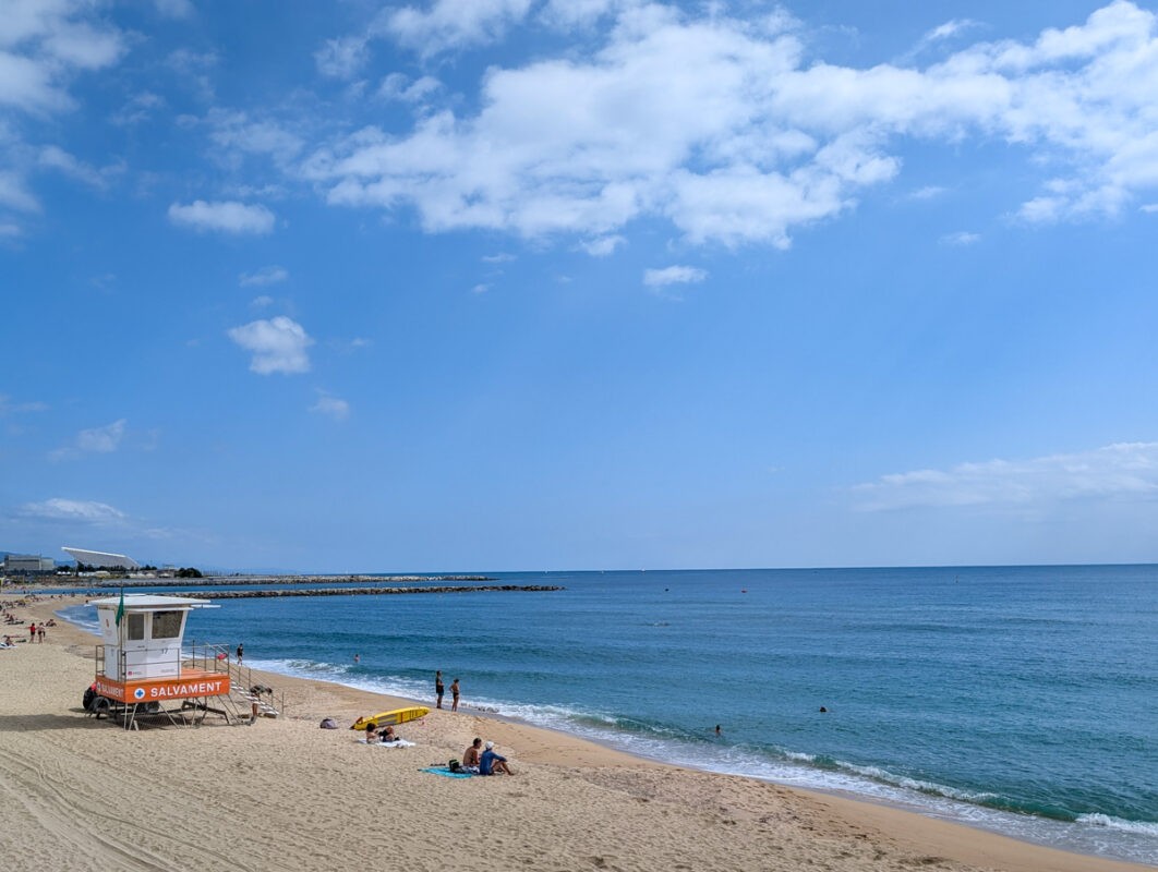 A peaceful beach scene with a lifeguard station labeled "Salvament" on the sand. The calm blue ocean stretches to the horizon under a clear sky, with a few people sunbathing and swimming.