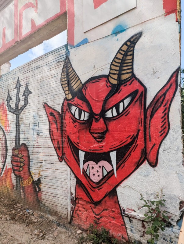 A vibrant street mural depicting a red demon with horns, sharp fangs, and exaggerated features. The demon holds a pitchfork, and the graffiti is painted on a white, textured wall.