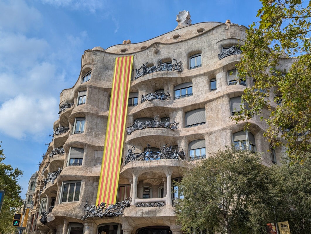 A view of Casa Milà, also known as La Pedrera, in Barcelona. The modernist building features a unique undulating stone facade and is adorned with a large vertical Catalan flag hanging from the top. Sculptural elements decorate the balconies.