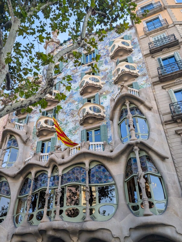 A close-up of Casa Batlló in Barcelona, designed by Antoni Gaudí. The building’s ornate facade includes organic, bone-like shapes, vibrant mosaic tiles, and curved windows. A Catalan flag is also visible, hanging from a balcony.